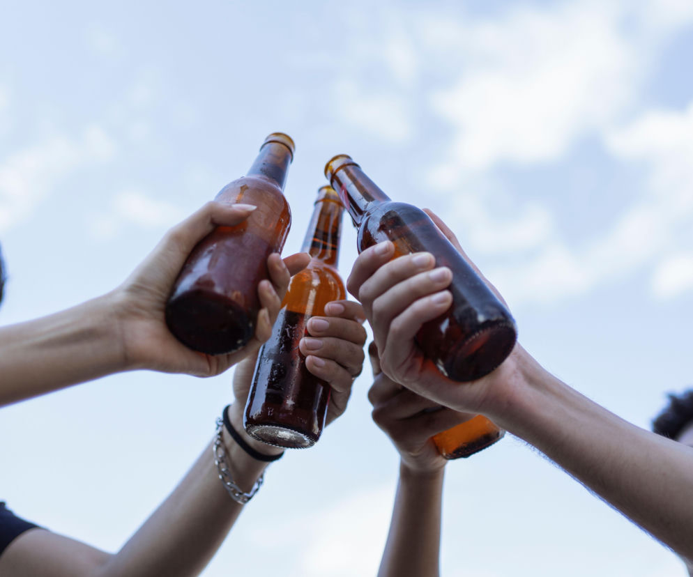 Dłonie osób wznoszących toast butelkami piwa na tle nieba. O popularności piwa 0% w Polsce przeczytasz w Eska.