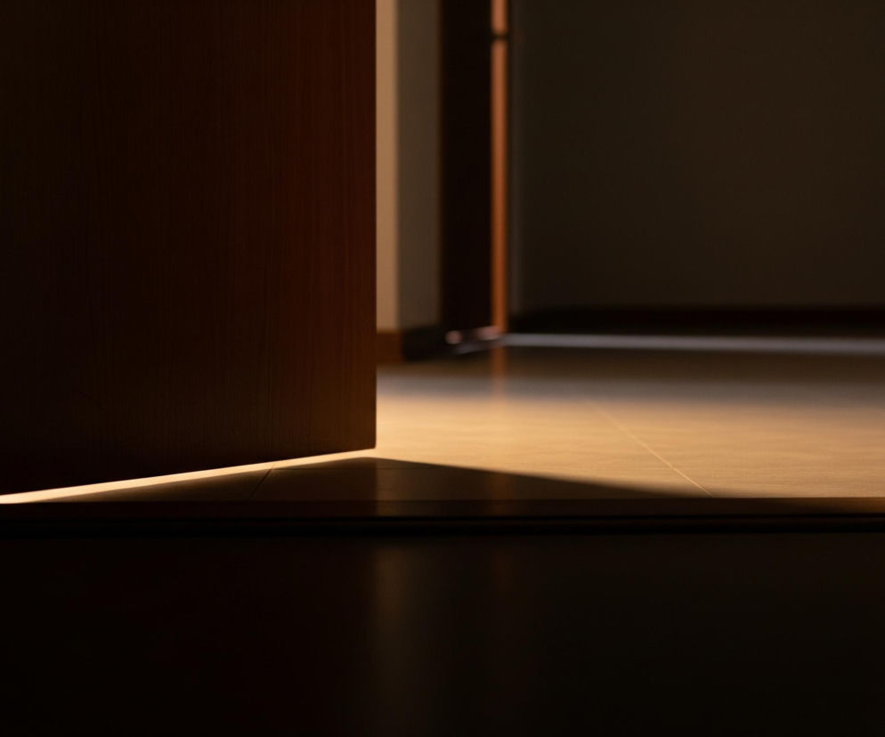 Częściowo otwarte, ciemnobrązowe drzwi uchylają się w prawą stronę, ukazując jasne, ciepłe światło padające na podłogę. Światło tworzy ostry trójkątny cień na ciemnym panelu podłogowym w pierwszym planie. Za otwartymi drzwiami widać kolejny, równie ciemny, odleglejszy pokój, rozjaśniony ciepłym, beżowym światłem na podłodze, z linią cienia biegnącą wzdłuż listwy przypodłogowej.