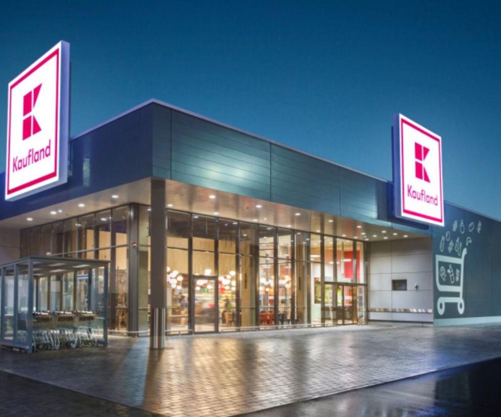 Nowoczesny budynek supermarketu Kaufland wieczorem. Nad wejściem dwa podświetlone logotypy sieci handlowej. Przed budynkiem przeszklona wiata na wózki sklepowe i mokry chodnik po deszczu. O kwestiach podwyżek w Kauflandzie przeczytasz na Super Biznes.