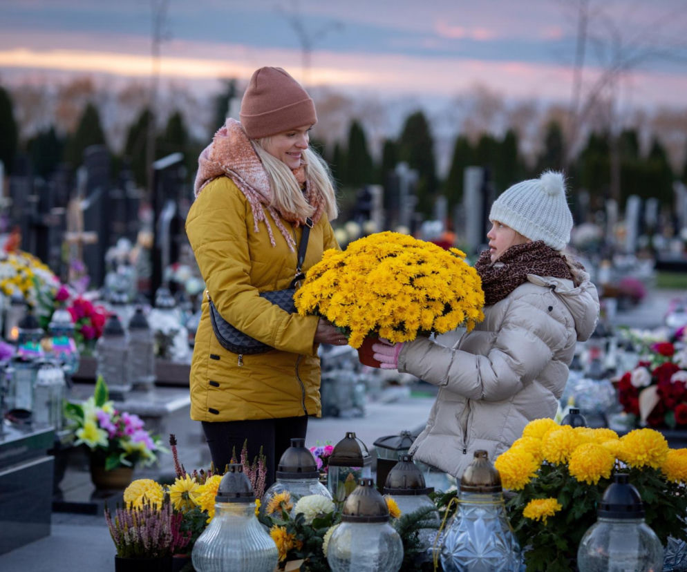 Kobieta i dziewczynka trzymają żółte chryzantemy na cmentarzu. O przesądach na 1 listopada przeczytasz na SE.