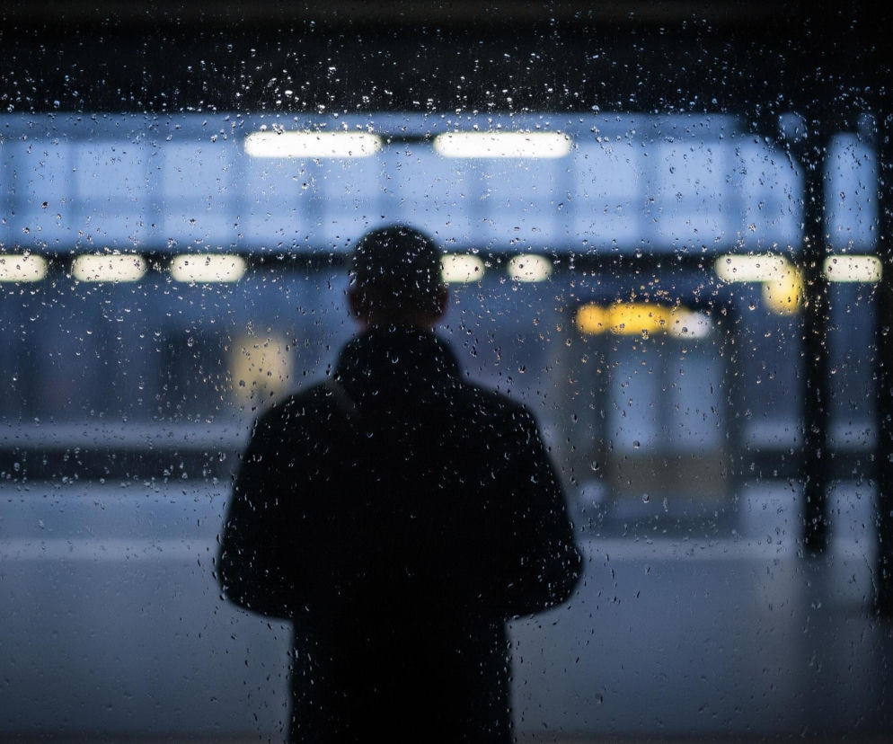 Centralnym elementem jest sylwetka osoby, widoczna od tyłu, ubrana w ciemną kurtkę, stojąca za szybą pokrytą licznymi kroplami wody. Tło przedstawia rozmyty widok dworca lub stacji metra z rzędami jasnych, prostokątnych świateł odbijających się w mokrej nawierzchni. Dominują chłodne odcienie niebieskiego i szarości, z kilkoma żółtymi akcentami świateł w oddali, tworząc melancholijny nastrój. Krople wody na szybie rozpraszają światło, dodając zdjęciu głębi i tekstury.