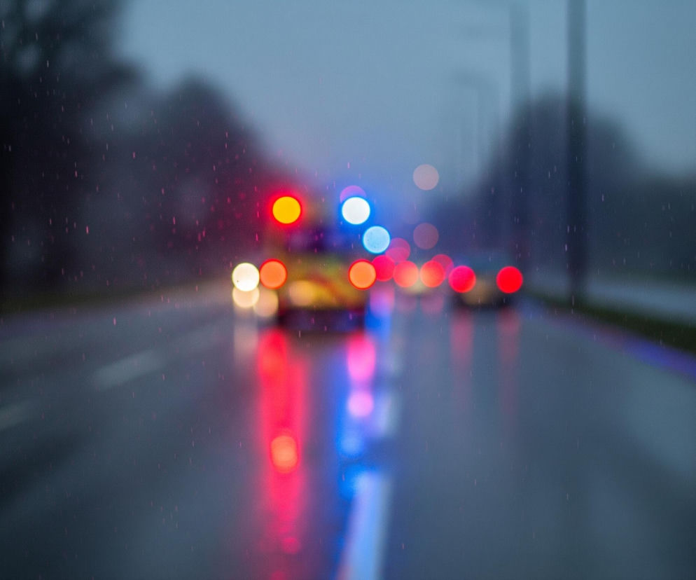 Rozmyte światła pojazdów ratunkowych, w tym czerwone, niebieskie i żółte, dominują centralną część obrazu, odbijając się na mokrej nawierzchni drogi. W górnej części widoczne są niewyraźne sylwetki drzew i ciemnego nieba, natomiast na całej powierzchni zdjęcia rozproszone są drobne, jasne punkciki, imitujące krople deszczu lub śniegu. Białe i niebieskie linie wyznaczają pasy ruchu na ciemnym asfalcie, a po prawej stronie majaczy rozmazany znak drogowy.