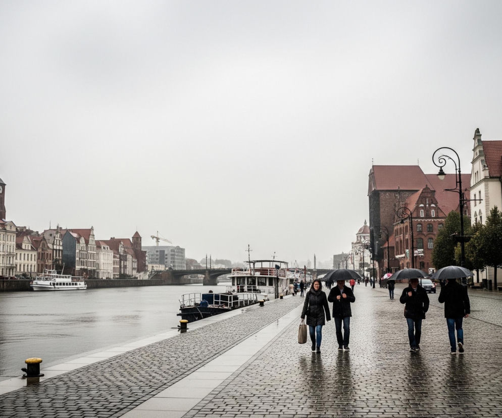 Cztery osoby idą chodnikiem z bruku, trzy z nich niosą otwarte czarne parasole, a pierwsza kobieta po lewej trzyma torbę na zakupy. Po prawej stronie chodnika rosną młode drzewa, a dalej widać zabytkowe kamienice z czerwonymi dachami i jasnymi fasadami, z których niektóre mają balkony. Po lewej stronie widać rzekę z kilkoma zacumowanymi łodziami i statkami wycieczkowymi, a w oddali most i wysoką wieżę kościelną z zegarem i szpiczastym dachem. Szare, zachmurzone niebo dominuje nad horyzontem, a mokry chodnik i rzeka odbijają światło.