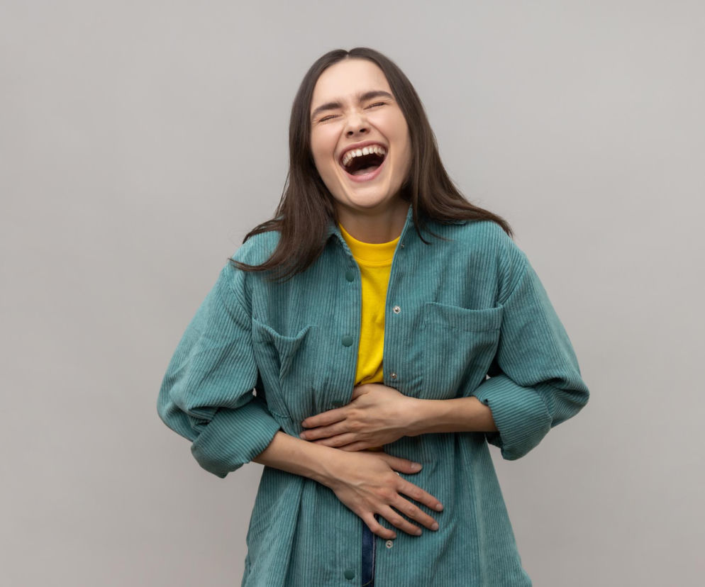 Młoda kobieta w żółtej koszulce i zielonej koszuli w sztruksy, śmiejąca się z zamkniętymi oczami i otwartymi ustami, trzymająca się za brzuch, prawdopodobnie z powodu kolki śmiechowej. Na Poradnik Zdrowie znajdziesz sposoby na ten problem.