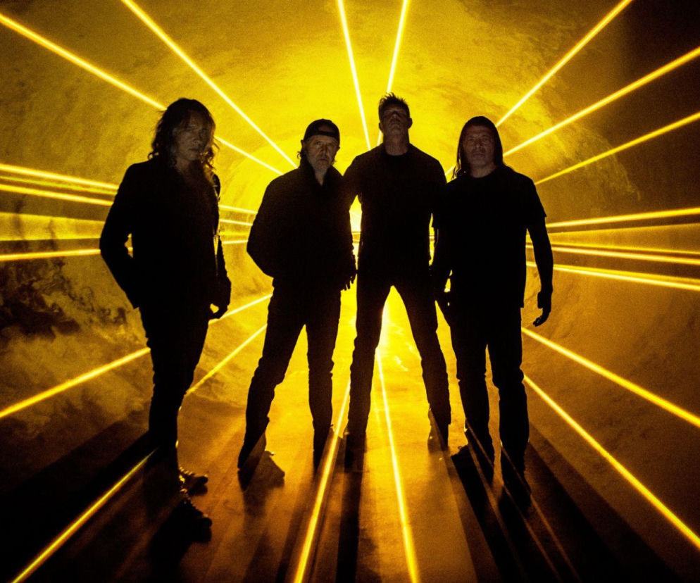 Czworo muzyków zespołu Metallica na tle żółtych wiązek światła. O fanach grupy przeczytasz na EskaRock.