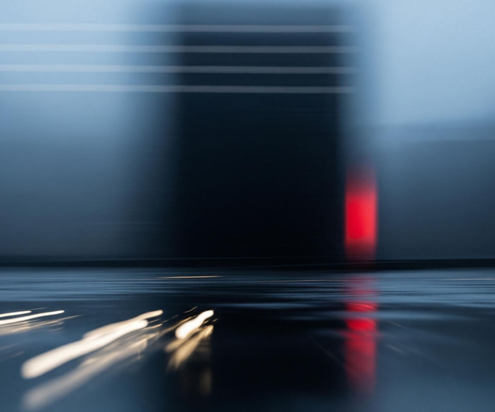 Abstrakcyjną kompozycję z rozmytymi kształtami i smugami światła. W tle widoczny jest pionowy, ciemny prostokąt, a po jego prawej stronie pionowy, intensywnie czerwony pasek, oba rozmazane horyzontalnie. Całość tła utrzymana jest w odcieniach szarości i błękitu, z kilkoma poziomymi, jasnymi liniami w górnej części. Na pierwszym planie znajduje się błyszcząca, ciemna powierzchnia odbijająca światło, z horyzontalnymi, białymi i żółtymi smugami, które rozciągają się w głąb obrazu. Czerwone światło z tła również odbija się na tej powierzchni, tworząc pionowy, rozmyty pas.