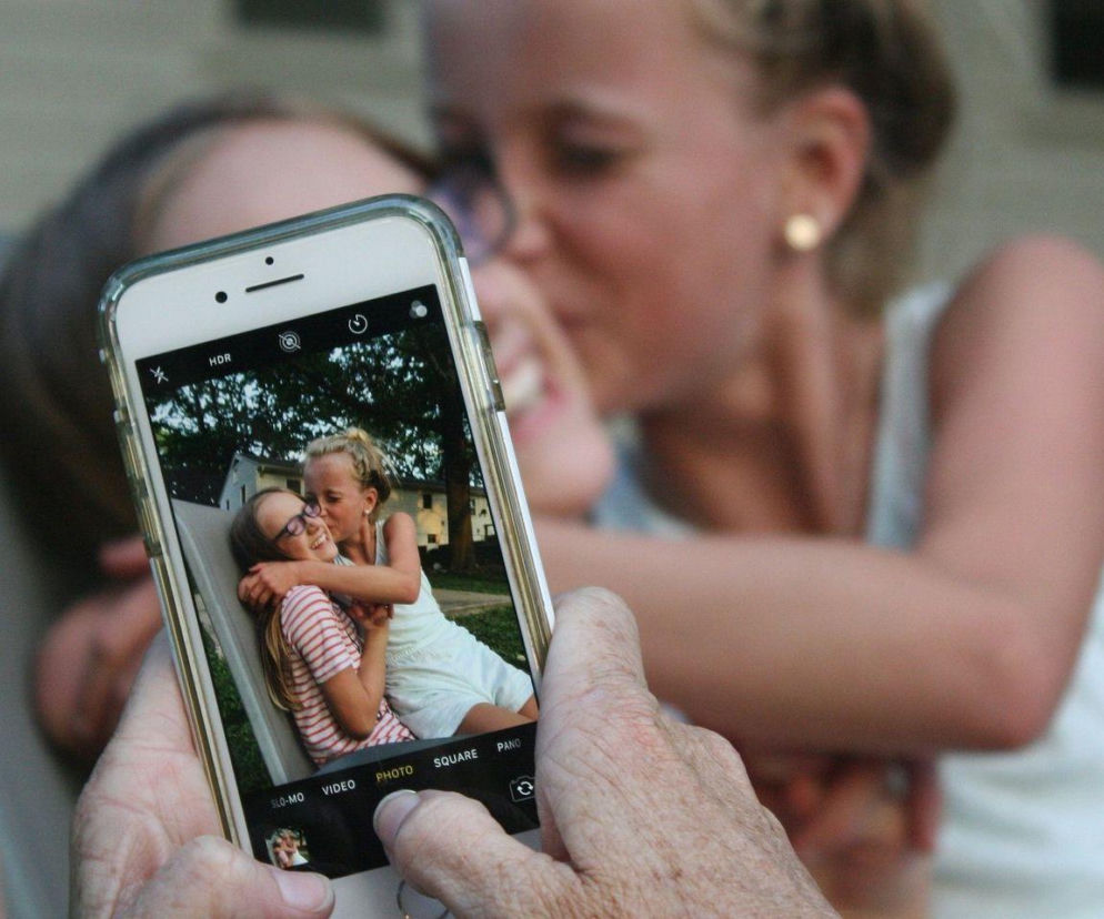 Dłonie osoby fotografującej smartfonem dwie przytulone dziewczynki. O cyfrowej etykiecie przeczytasz w serwisie Eska.