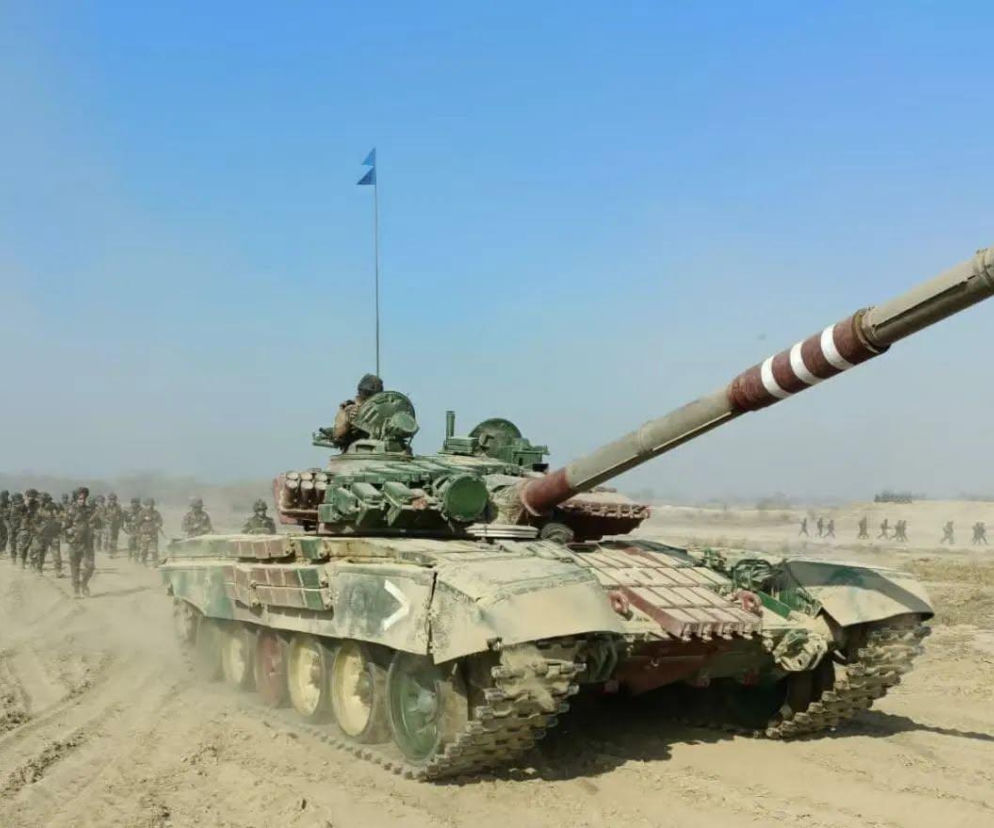 Indyjski czołg T-72 w pustynnym kamuflażu na poligonie, w tle idą żołnierze. Armia Indii planuje przekształcić te czołgi w bezzałogowe platformy, o czym przeczytasz więcej na naszym portalu.