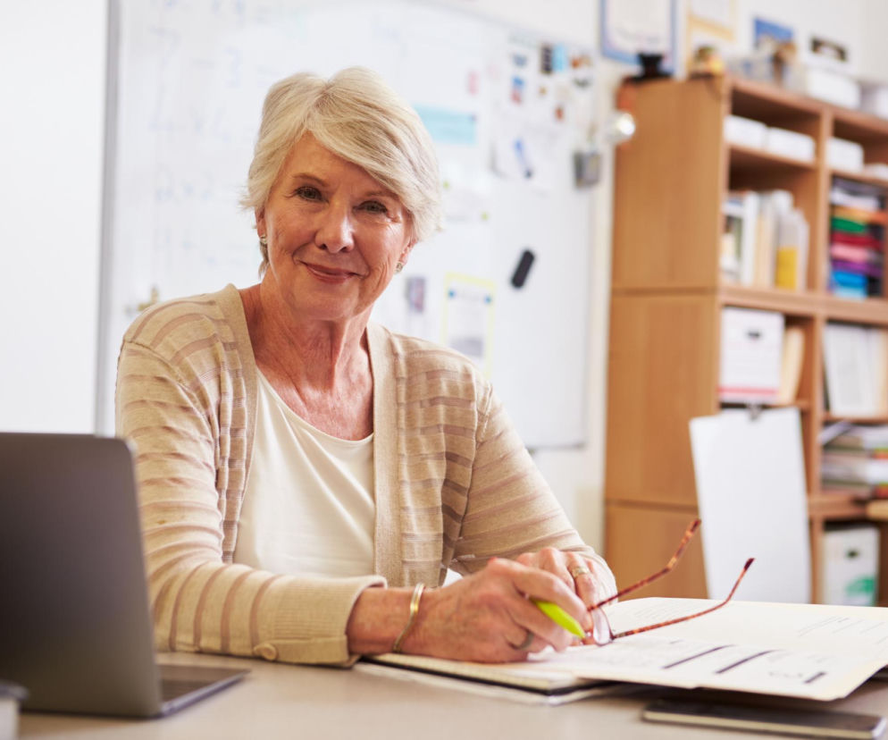 Siedząca przy biurku starsza kobieta o siwych włosach, ubrana w białą bluzkę i beżowy sweter, uśmiecha się, trzymając okulary. Przed nią laptop i dokumenty, a w tle tablica i regał z segregatorami. Temat dorabiania do emerytury omawia portal Super Biznes.