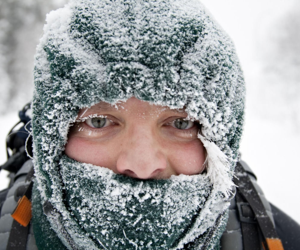 Twarz osoby zmarzniętej w zielonej czapce i szaliku pokrytych szronem, z widocznymi tylko oczami, rzęsami i brwiami obsypanymi śniegiem, na tle zimowego pejzażu. Sprawdź, jak chronić się przed mrozem na Poradnik Zdrowie.