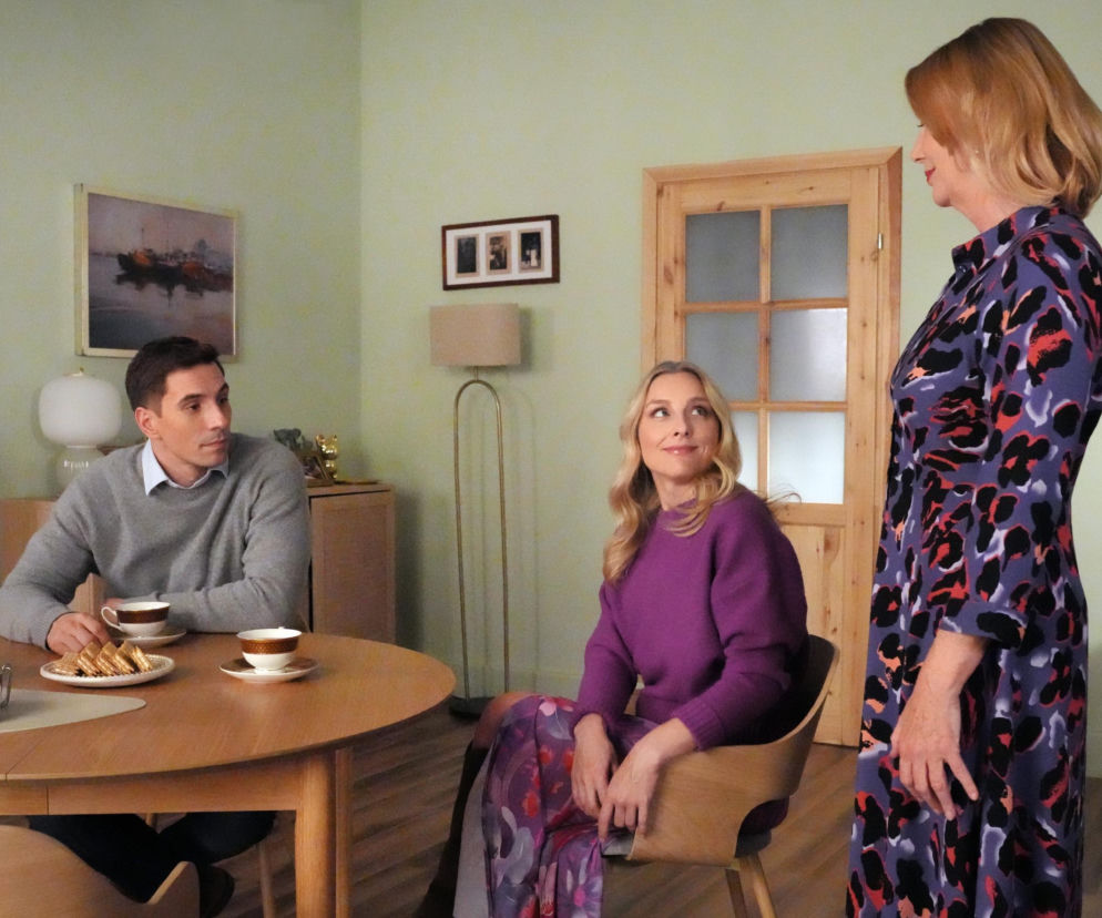 Trzy osoby w pomieszczeniu, Wanda (Maja Barełkowska) w kolorowej sukience z nadrukiem stoi po prawej, uśmiechnięta kobieta w fioletowym swetrze siedzi pośrodku, a mężczyzna w szarym swetrze przy stole z kawą po lewej. Nowe życie Wandy i bliskość Jerzego w Barwach szczęścia to główny temat na Super Seriale.