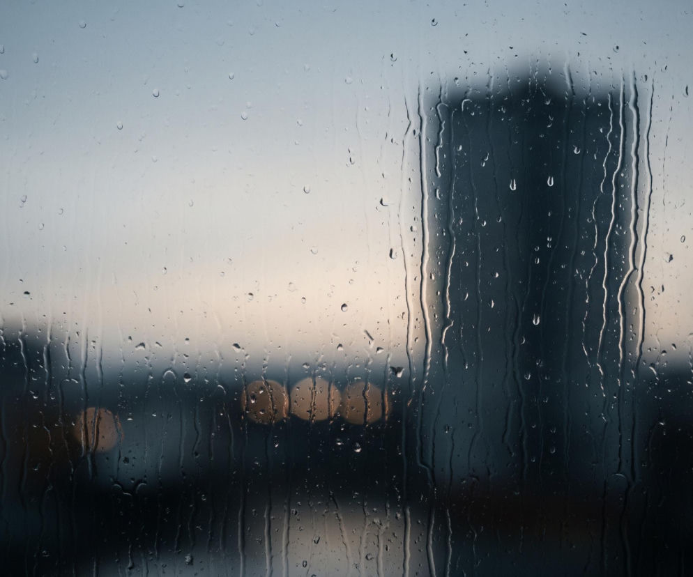 Szyba pokryta jest licznymi kroplami deszczu o różnych rozmiarach i kształtach, a także smugami wody spływającymi pionowo w dół. Za mokrą szybą rozmywa się miejski krajobraz, dominują ciemne, nieostre sylwetki budynków, w tym jeden wyraźnie większy i pionowy, położony w prawej części kadru. W tle widać również kilka rozmytych, okrągłych punktów świetlnych o ciepłej, pomarańczowej barwie, przypominających światła uliczne lub okna. Górna część nieba ma odcień chłodnego błękitu przechodzącego w jaśniejszy, lekko różowawy ton bliżej horyzontu.