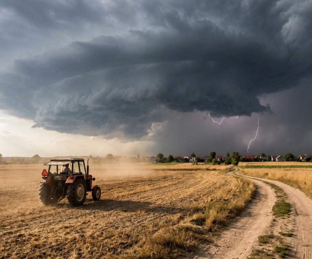 Czerwony traktor na zaoranym polu, z widocznym błękitnym niebem po lewej i potężną, ciemną chmurą burzową typu shelf cloud z błyskawicami po prawej stronie. Z daleka widać wiejskie domy, a obok pola prowadzi polna droga. Ostrzeżenia o burzach i upałach przeczytasz na naszym portalu.