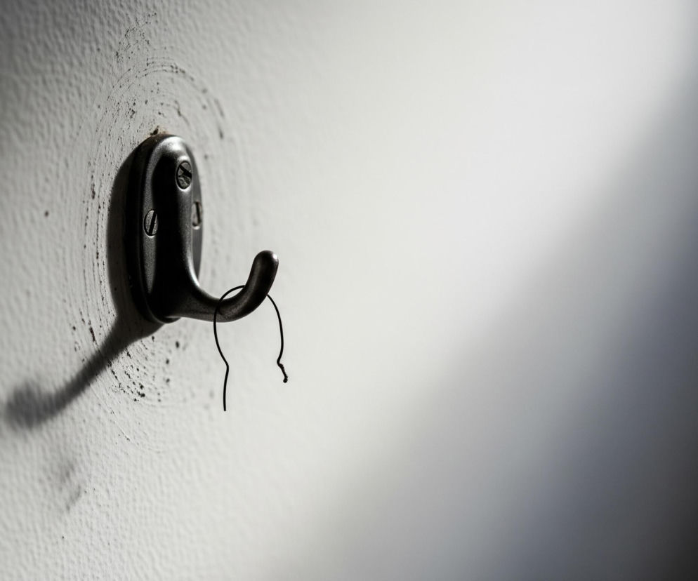 Na zdjęciu centralnie znajduje się ciemny, metalowy haczyk, przymocowany do jasnej, chropowatej ściany dwoma śrubami z płaskim nacięciem. Pod haczykiem wisi cienki, ciemny, wygięty drut. Obszar wokół haczyka jest wyraźnie zabrudzony, z widocznymi ciemnymi smugami i kropkami, kontrastującymi z ogólnym jasnym kolorem ściany. Silne oświetlenie z lewej strony tworzy wyraźny cień rzucany przez haczyk i drut, który rozciąga się w lewo i w dół, a prawa strona kadru jest znacznie ciemniejsza, przechodząc w głęboką czerń.