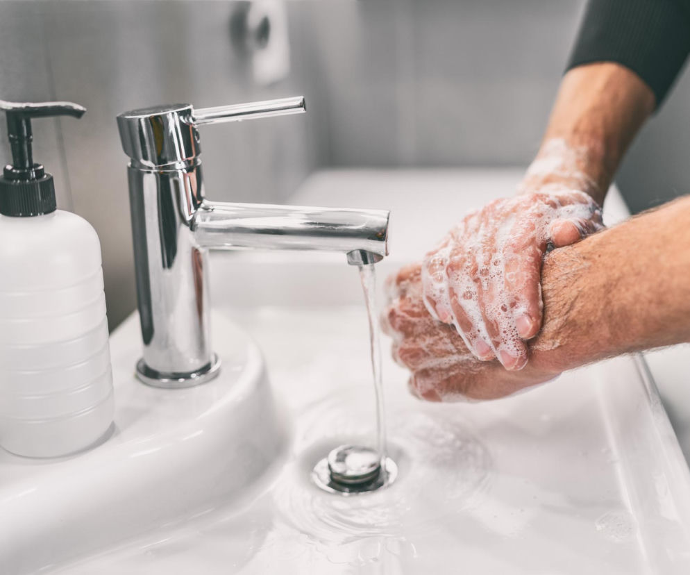 Osoba myjąca ręce mydłem pod kranem w umywalce, piana pokrywa dłonie, co symbolizuje prawidłową higienę. Artykuł na Poradnik Zdrowie wyjaśnia, jak skutecznie myć ręce, aby chronić skórę.