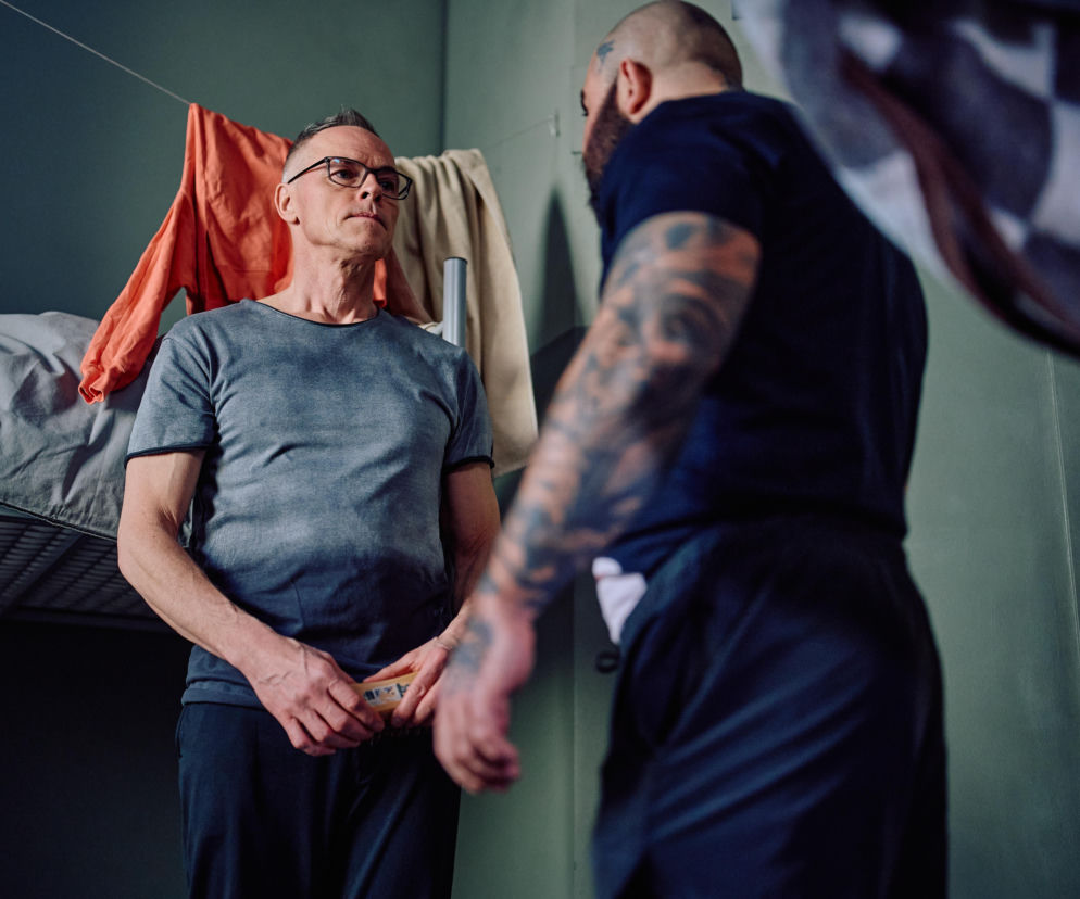 Mężczyzna w okularach i szarej koszulce rozmawia z brodatym mężczyzną z tatuażami w ciemnej koszulce, stojąc w celi więziennej. Na tle widać piętrowe łóżko i wiszące ubrania, co podkreśla trudną sytuację Damiana z Na Wspólnej. Więcej o tym na portalu Super Seriale.