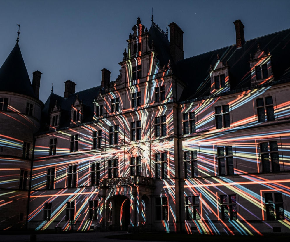 Duży, zabytkowy budynek z wieloma oknami jest oświetlony kolorowymi, dynamicznymi promieniami świetlnymi w odcieniach czerwieni, pomarańczy, żółci, zieleni i błękitu, które rozchodzą się od centralnych punktów. Budowla ma jasną fasadę z ciemnymi, spadzistymi dachami i licznymi kominami, a po lewej stronie dominuje okrągła wieża z dachem stożkowym. Na tle ciemnego, wieczornego nieba widoczne są fragmenty nagich drzew po lewej stronie oraz ledwo dostrzegalne gwiazdy.