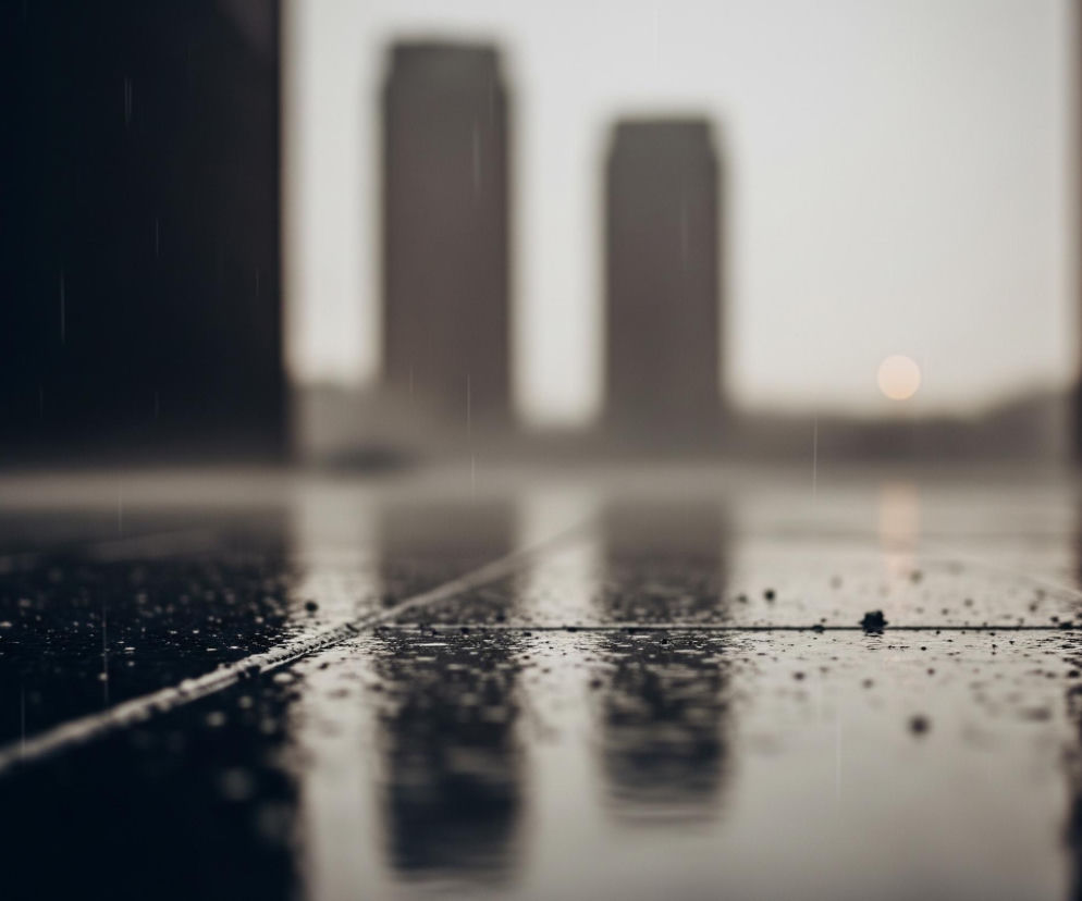 Błyszcząca, ciemna, mokra posadzka z widocznymi kroplami deszczu zajmuje pierwszy plan, ukazując wyraźne linie podziału między płytami. W tle, rozmyte sylwetki dwóch wysokich budynków w szarych odcieniach dominują centralną część, a na horyzoncie widoczna jest jasna, okrągła plama, prawdopodobnie słońce lub księżyc. Po lewej i prawej stronie kadru, z ciemnych, pionowych powierzchni padają długie, rozmyte cienie, a w całym obrazie unoszą się liczne, cienkie smugi deszczu.