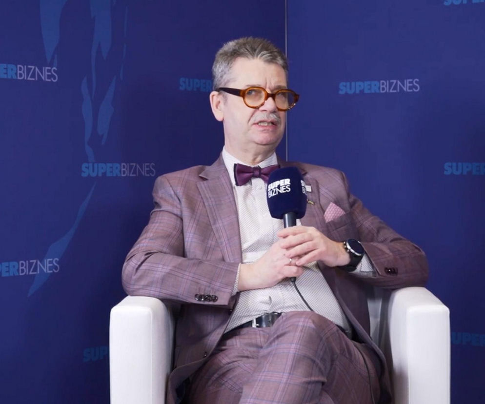 Prof. Piotr Wachowiak, rektor SGH, siedzący w fioletowym garniturze w kratę i brązowych okularach, trzyma mikrofon z logo Super Biznes. Tło stanowi niebieska ściana z powtarzającym się napisem Super Biznes. O stanie polskiej gospodarki można przeczytać na portalu Super Biznes.