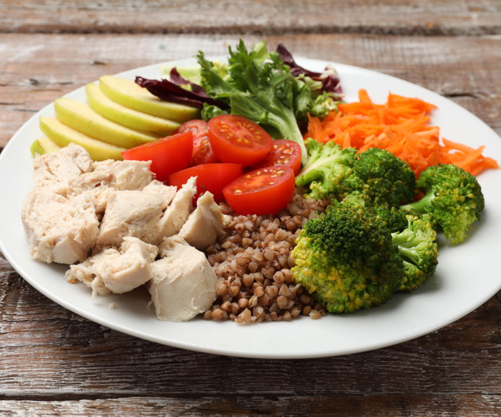 Talerz z gotowanym kurczakiem, kaszą gryczaną, kawałkami jabłka, pomidorami, brokułami oraz tartą marchewką i sałatą, symbolizujący zbilansowaną dietę. Więcej o zdrowym odżywianiu i jego wpływie na zdrowie przeczytasz na Poradnik Zdrowie.