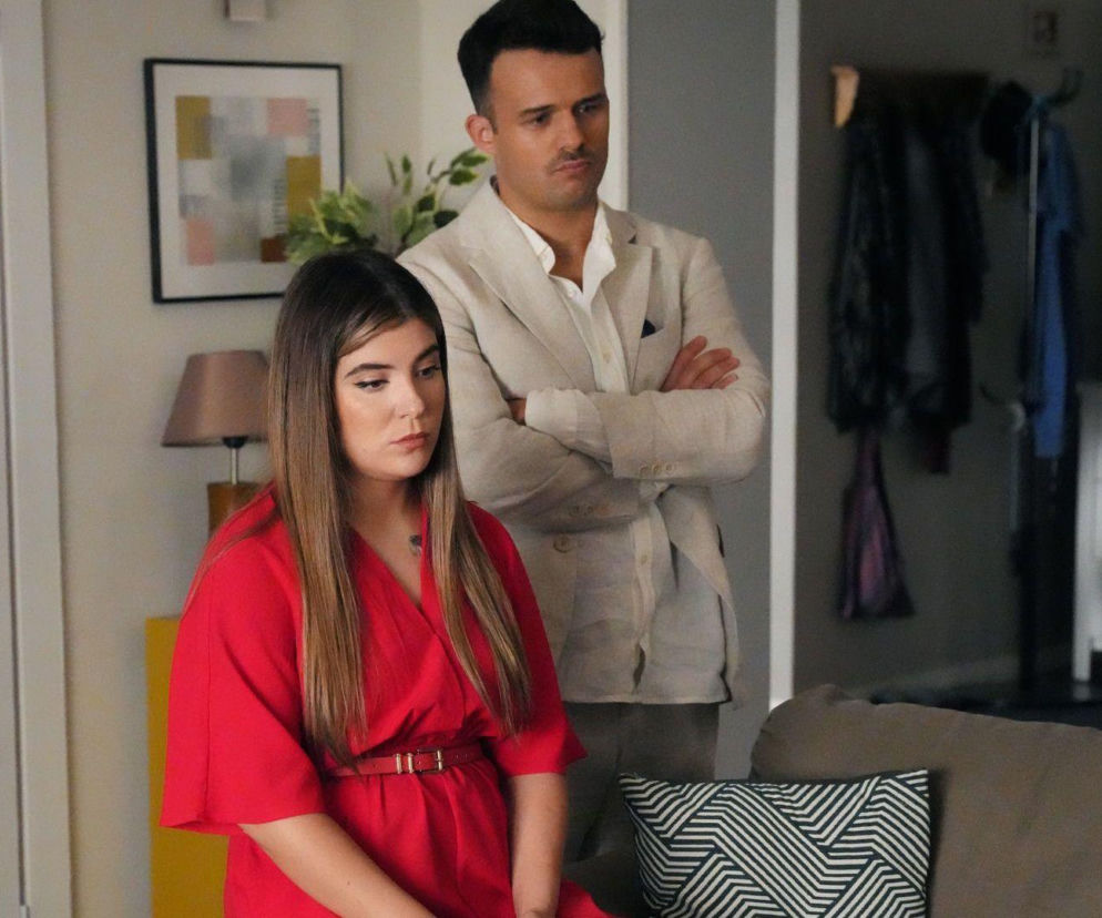 Agata (Natalia Zambrzycka) w czerwonej sukience i Ignacy (Olaf Staszkiewicz) w beżowej marynarce. Mężczyzna ma skrzyżowane ramiona i poważną minę, kobieta patrzy w dół. Odcinek 3352 serialu Barwy szczęścia dostępny na Super Seriale.