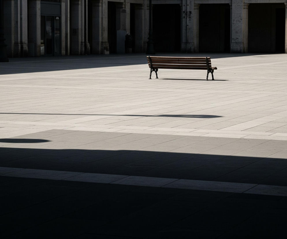 Drewniana ławka z ciemnymi, metalowymi nóżkami stoi na środku obszernego placu wyłożonego jasnoszarymi płytami. Po prawej stronie ławki widoczne są schody wchodzące na podwyższenie, a w tle rozciąga się rządek kamiennych kolumn i ciemnych arkad, rzucających głębokie cienie na plac. Na pierwszym planie i po lewej stronie placu dominuje duża, ciemna plama cienia, a od ławki w prawo i w dół rozciąga się długi, wąski cień.