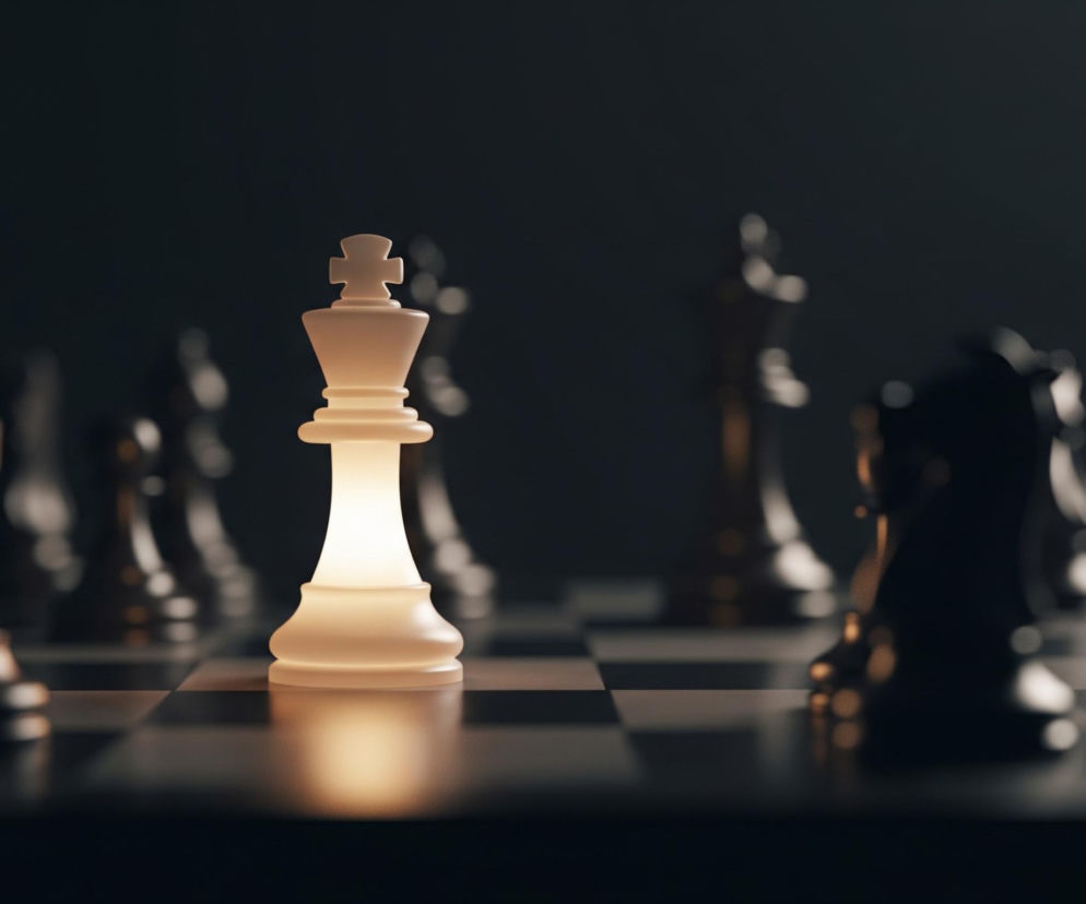 Na pierwszym planie, centralnie, stoi świecący na kremowo biało król szachowy. Figura wydziela ciepłe, bursztynowe światło, które rozjaśnia jej podstawę i tworzy rozmyte odbicie na czarno-białej szachownicy. Wokół króla, w półmroku, rozłożone są inne ciemne figury szachowe, większość z nich jest niewyraźna, co podkreśla skupienie na centralnej postaci. Całość spowita jest ciemnym tłem, z lekkim gradientem światła w górnej części.