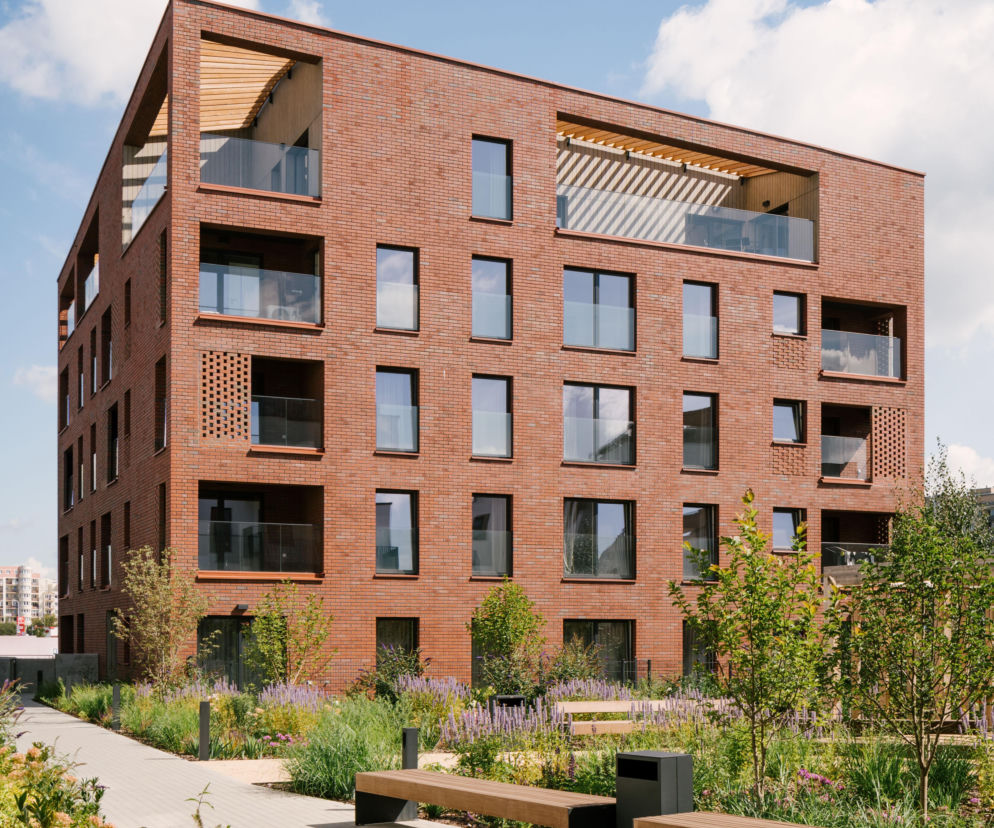 Nowoczesny budynek Nordic Bemowo z ceglaną fasadą, balkonami z przeszkleniami i drewnianymi akcentami, otoczony zielenią i ścieżkami z ławkami. Projekt pracowni Kuryłowicz & Associates z Warszawy zgłoszony do konkursu Życie w Architekturze 2025. Dowiedz się więcej na Architektura Murator Plus.