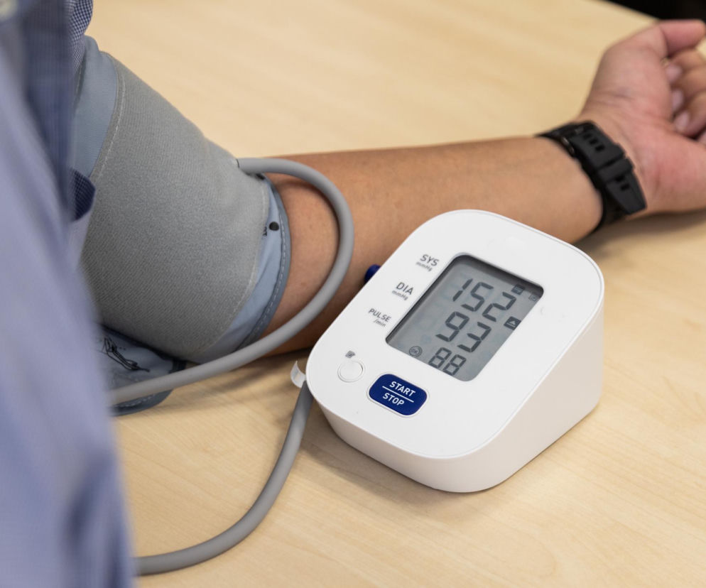 Osoba mierząca ciśnienie krwi automatycznym ciśnieniomierzem naramiennym; na wyświetlaczu widoczne wysokie wartości 152/93. O tym, jak obniżyć wysokie ciśnienie krwi i kontrolować nadciśnienie tętnicze, przeczytasz na Poradniku Zdrowie.