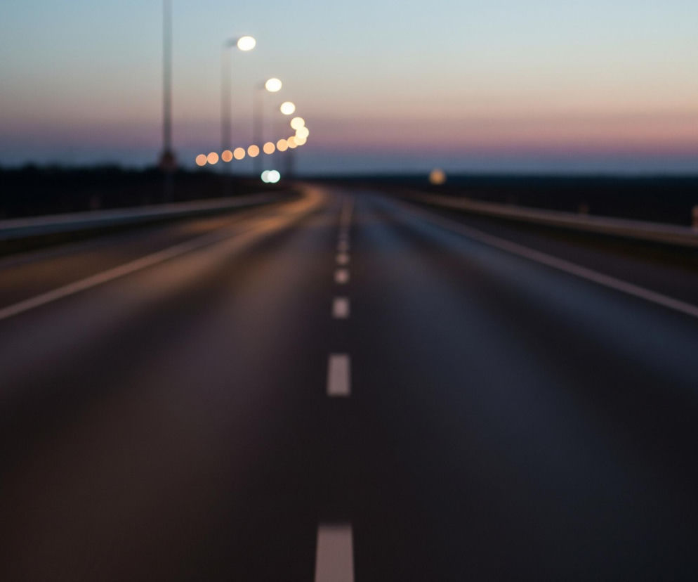 Rozmyty widok pustej drogi asfaltowej w nocy lub o zmierzchu, z liniami przerywanymi na środku. Po obu stronach drogi znajdują się bariery ochronne, a w oddali widać szereg rozmytych, kulistych świateł ulicznych, które tworzą łuk wzdłuż zakręcającej drogi. Horyzont prezentuje gradient barw od ciemnego granatu u dołu do jasnego błękitu na samej górze, przechodząc przez odcienie różu i fioletu.