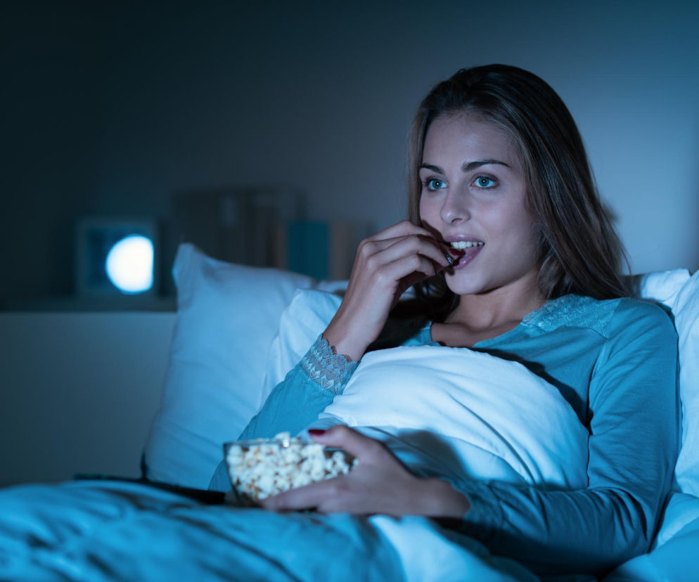 Młoda kobieta w piżamie, leżąca w łóżku i jedząca popcorn podczas oglądania filmu w ciemnym pokoju, symbolizująca nocny tryb życia. Na Poradnik Zdrowie można przeczytać o związku późnego chodzenia spać z ryzykiem udaru.