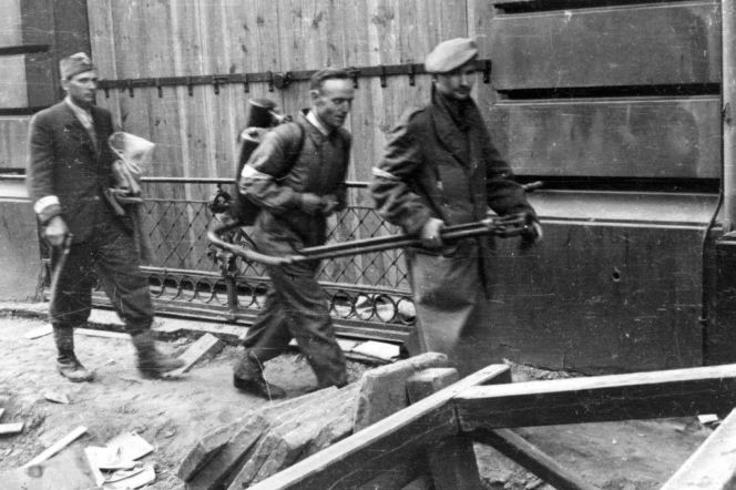 Trzej powstańcy warszawscy z bronią idą obok barykady. O tym, ilu bohaterów wciąż żyje, przeczytasz na Eska Warszawa.