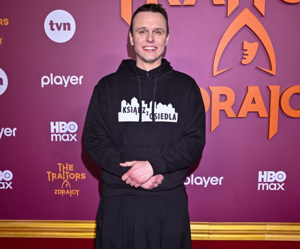 Rafał Główczyński, znany jako Ksiądz z osiedla, uśmiechnięty na ściance promocyjnej programu The Traitors. Zdrajcy. Ma na sobie czarną bluzę z napisem Ksiądz z osiedla. Obok widoczne są loga TVN i HBO Max, a także fragmenty nazwy programu, o którym możesz przeczytać na Super Seriale.