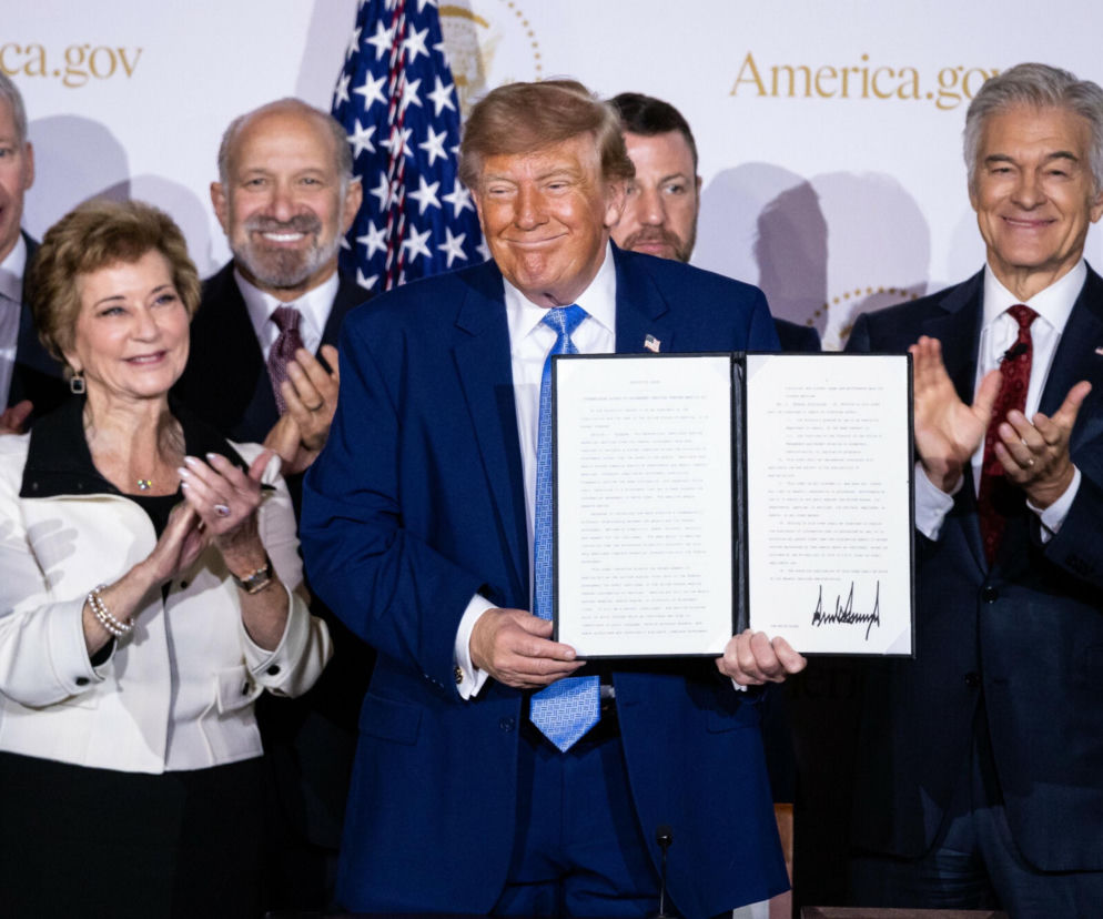Donald Trump prezentuje podpisany dokument. Wokół stoją uśmiechnięte osoby. O konstytucji dla AI pisze SE Superbiz.