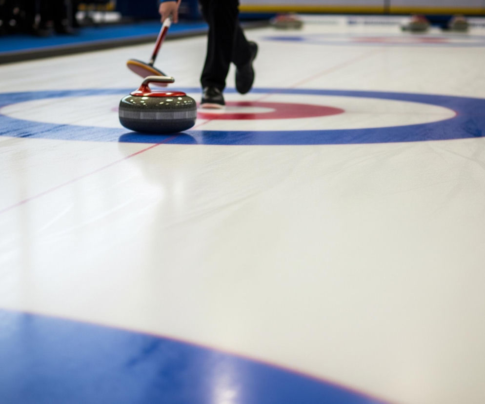 Na błyszczącej, białej tafli lodu widoczny jest kamień do curlingu z czerwonym uchwytem i ciemnym korpusem z jasnymi paskami, właśnie wypuszczany przez osobę w czarnych spodniach i butach. Postać ta, częściowo widoczna od pasa w dół, trzyma kij do curlingu z czerwoną rączką i czarną końcówką. Na tafli lodu namalowane są koncentryczne okręgi w kolorach czerwonym i niebieskim, stanowiące cel. W tle, na lewej krawędzi obrazu, widać niewyraźne sylwetki innych ludzi i sprzętu, a w oddali kolejne kamienie do curlingu na podobnych polach gry.