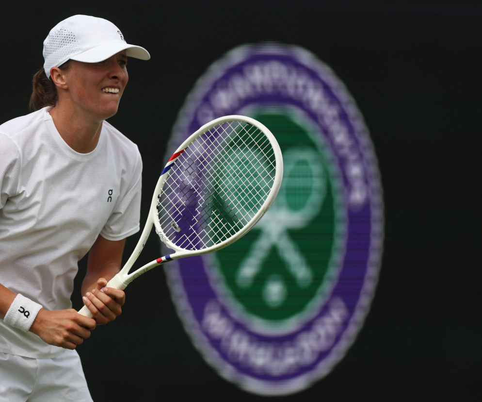 Tenisistka Iga Świątek w białym stroju sportowym, czapce i z rakietą, uśmiecha się na korcie Wimbledonu, przygotowując się do meczu z Taylor Townsend. O starcie Polki w turnieju przeczytasz więcej na naszym portalu.