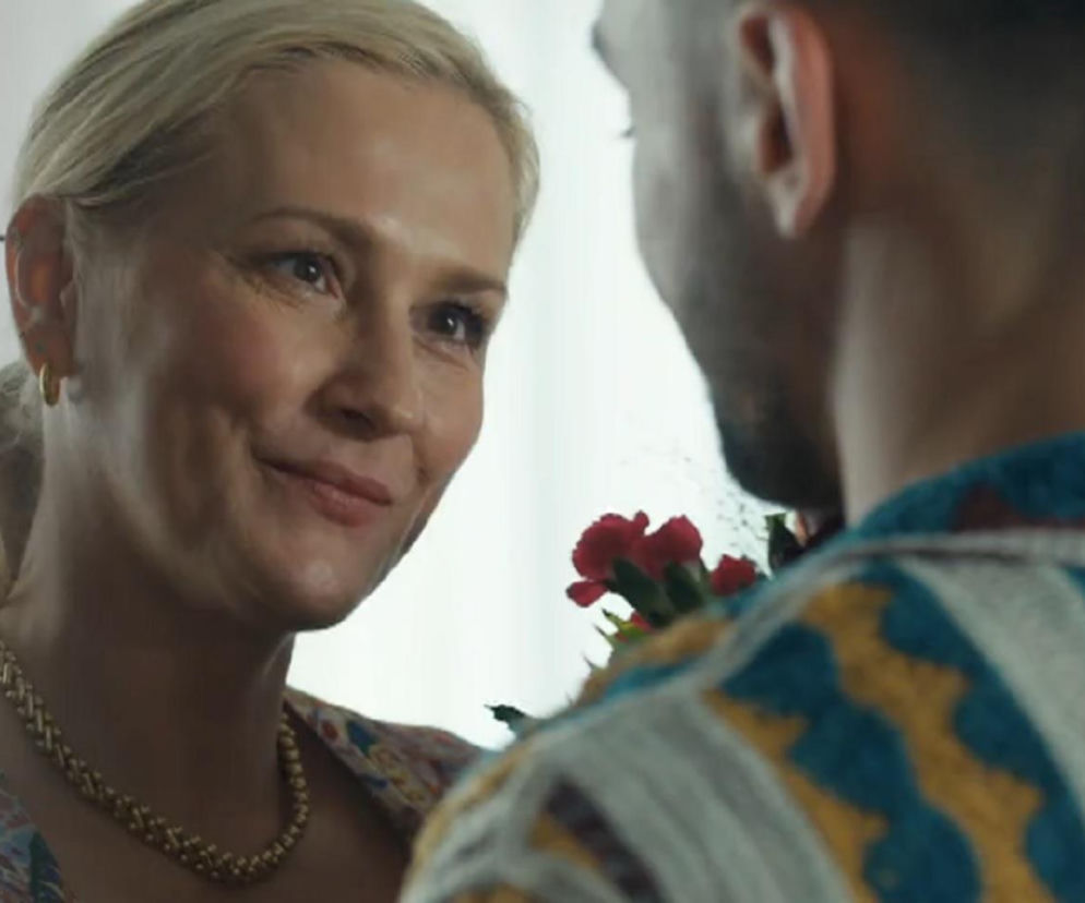 Uśmiechnięta Justyna (Sylwia Juszczak-Krawczyk) z blond włosami upiętymi w kucyk i złotym naszyjnikiem patrzy z boku na Mirka (Oliwier Kozłowski), który trzyma czerwone kwiaty. Scena z serialu Barwy szczęścia, której szczegóły znajdziesz na Super Seriale.