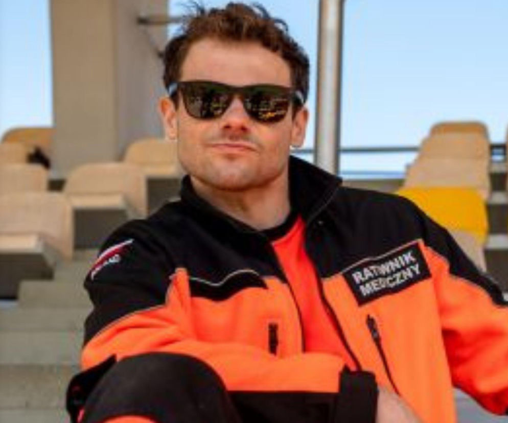 Mężczyzna w pomarańczowo-czarnym uniformie ratownika medycznego i okularach przeciwsłonecznych, siedzący na trybunach stadionu. Jest to kadr z serialu Na sygnale, który możesz obejrzeć i przeczytać o nim na Super Seriale.