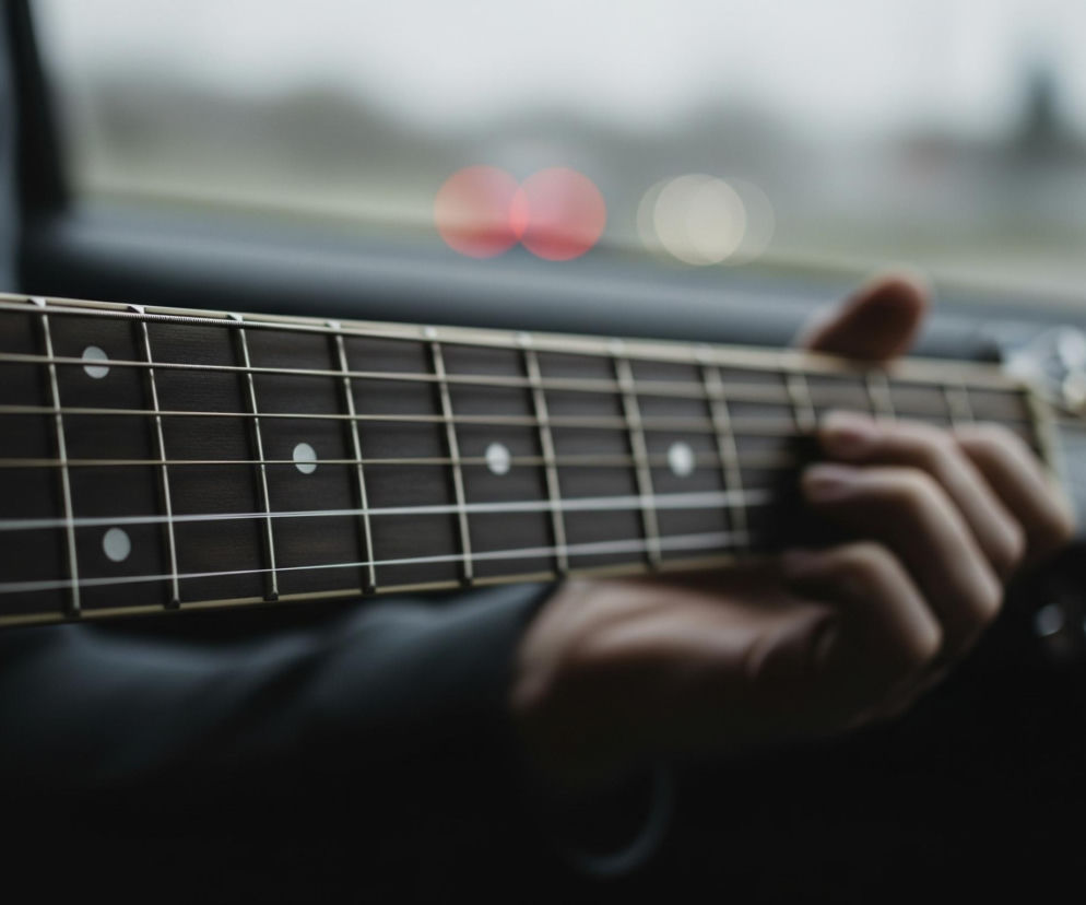 Mężczyzna trzyma gitarę akustyczną, a jego lewa dłoń, ubrana w ciemny rękaw, dociska struny do ciemnego gryfu. Na gryfie widać białe kropki pełniące funkcję markerów pozycji oraz metalowe progi. W tle, za gitarą i dłonią, rozmazane światła w kolorze czerwonym i białym, sugerują scenerię na zewnątrz, być może z pojazdu.