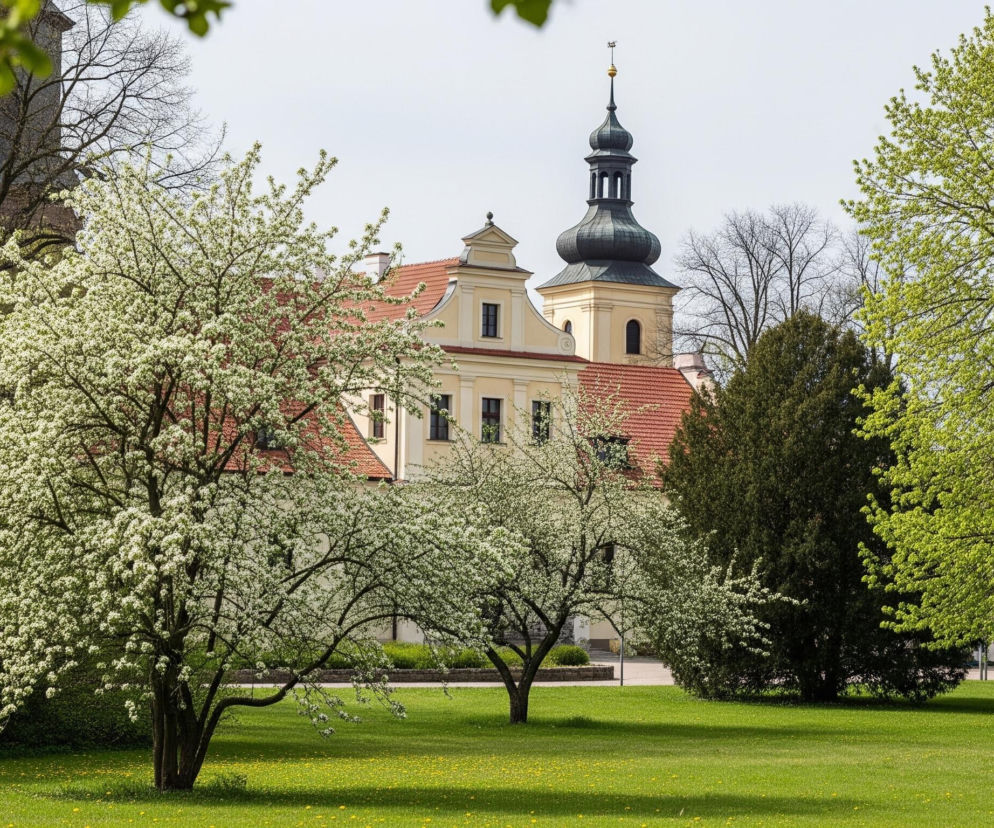 Na pierwszym planie, w centrum, znajduje się kwitnące drzewo o białych kwiatach, częściowo przysłaniające inne, również kwitnące drzewa oraz budynek za nim. Budynek ma jasne, kremowe ściany i czerwone dachy, a z jego prawej strony wystaje wieża z czarnym, cebulastym hełmem i złotą iglicą zakończoną wiatrowskazem. Cała scena osadzona jest na tle rozległego, zielonego trawnika usianego drobnymi, żółtymi kwiatami, otoczonego bujną zielenią drzew i krzewów o zróżnicowanych odcieniach zieleni, z których część jest jeszcze bezlistna.