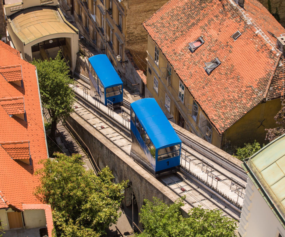 Dwa niebieskie wagoniki kolejki Uspinjača w Zagrzebiu między budynkami. O atrakcjach stolicy Chorwacji przeczytasz na SE.