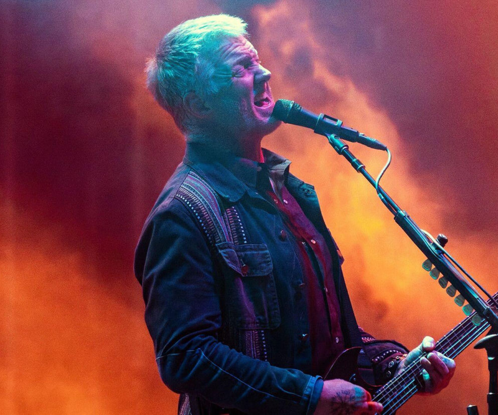 Josh Homme śpiewa do mikrofonu i gra na gitarze na scenie. O walce muzyka z rakiem przeczytasz na EskaRock.