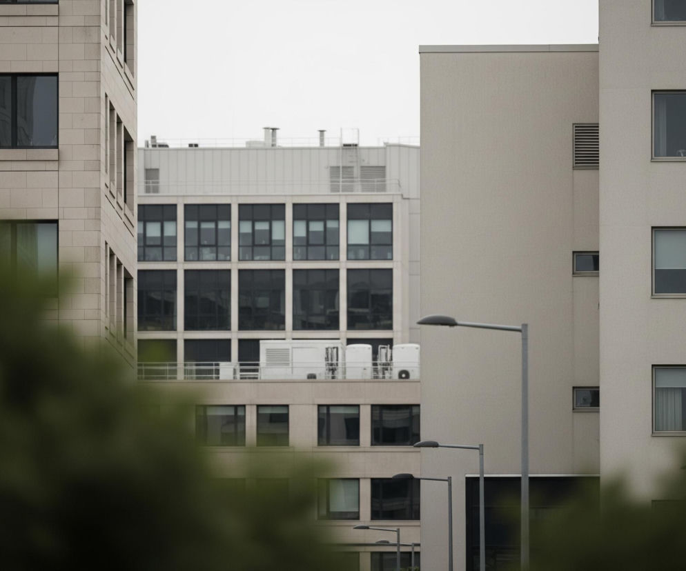 Po lewej stronie kadru znajduje się jasnobeżowy budynek z wyraźnie zaznaczonymi prostokątnymi oknami, niektóre z nich odbijają otoczenie. W tle, za nim, widoczny jest inny budynek z rzędami ciemnych okien i białymi konstrukcjami na dachu. Po prawej stronie kadru dominuje wysoki, jasnoszary budynek z mniejszymi oknami i poziomymi żaluzjami wentylacyjnymi. Na pierwszym planie, u dołu kadru, rozmyte zielone liście drzew częściowo zasłaniają widok, a pomiędzy budynkami widać dwie szare latarnie uliczne.