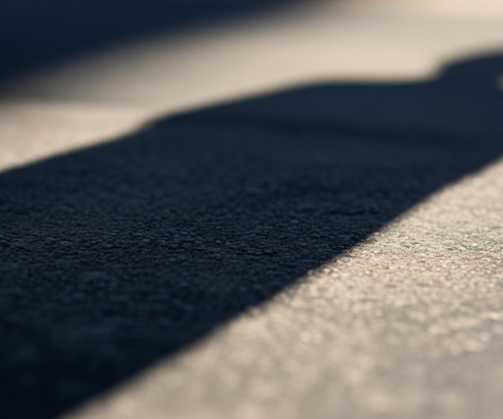 Ciemny cień o wydłużonym kształcie, przypominający sylwetkę człowieka z uniesioną głową, rozciąga się w poprzek piaszczystego, jasnoszarego podłoża. Na pierwszym planie cień jest ostro zarysowany i ma widoczną teksturę złożoną z drobnych, ciemnych ziaren, podczas gdy w oddali staje się coraz bardziej rozmyty. Światło padające z prawego górnego rogu tworzy wyraźną, jasną linię na granicy cienia i oświetlonej powierzchni.