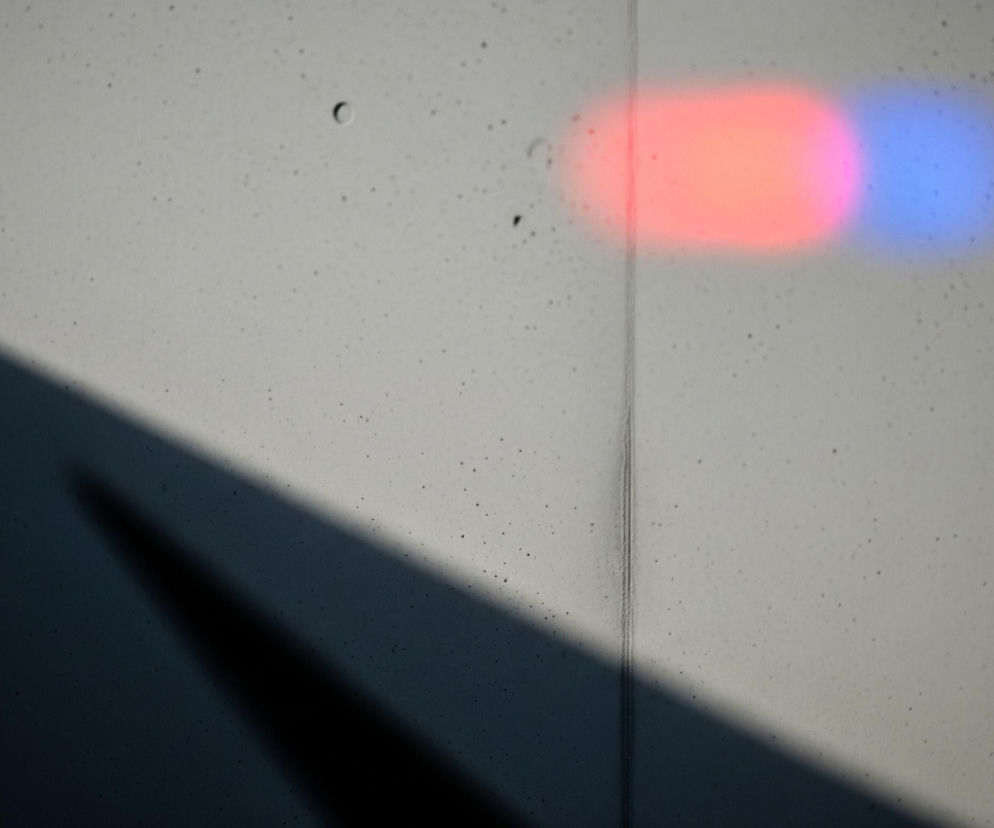 Na jasnoszarym, betonowym tle widoczne są dwa rozmyte, owalne kształty świetlne. Lewy kształt jest czerwony, a prawy niebieski, oba są wyraźnie jaśniejsze niż otoczenie. Po lewej stronie obrazu, od dolnego rogu do środkowej lewej krawędzi, rozciąga się ciemny cień, tworzący ukośną granicę z jaśniejszą częścią ściany. Pionowa linia, prawdopodobnie łączenie płyt, biegnie przez środek obrazu, oddzielając cień od światła i przechodząc przez prawą krawędź czerwonego światła. Cała powierzchnia betonu jest nieregularnie pokryta drobnymi, ciemnymi plamkami i kilkoma większymi, okrągłymi otworami.