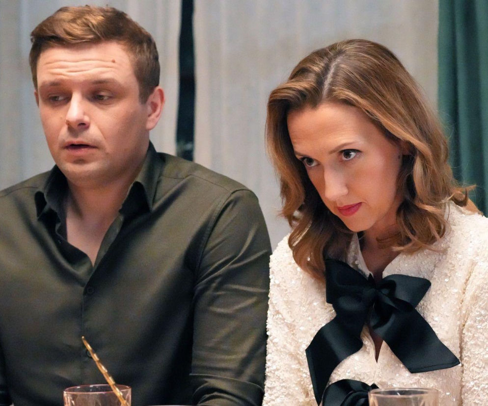 Anna Gzyra i Hubert Augustynowicz, aktorzy z serialu Barwy szczęścia, siedzący przy stole w stonowanych strojach. Kobieta ubrana w jasną, błyszczącą bluzkę z czarną kokardą, mężczyzna w ciemnozieloną koszulę. Więcej o ich rolach na portalu Super Seriale.