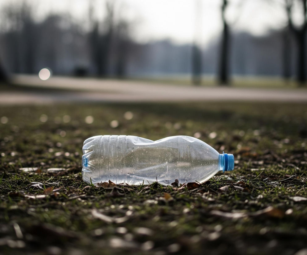 Na pierwszym planie leży plastikowa, przezroczysta butelka z niebieską nakrętką, zgnieciona w środkowej części. Butelka spoczywa na ciemnozielonej trawie, wśród której widoczne są brązowe, suche liście i drobne gałązki. Trawa jest częściowo oświetlona, tworząc obszary cienia. W tle rozciąga się niewyraźny krajobraz parku, z zamazanymi kształtami drzew o ciemnych pniach i jasnym, rozmytym niebem.