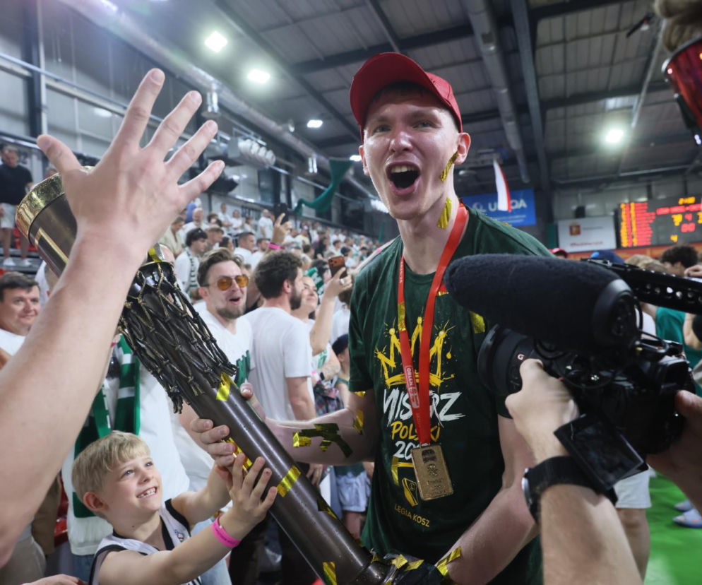 Koszykarz Andrzej Pluta w czerwonej czapce, z otwartymi ustami w okrzyku radości, z kawałkami konfetti na twarzy, trzyma puchar mistrzowski. Dziecko w koszulce Legii i inni kibice dotykają trofeum, a operator kamery filmuje scenę. Więcej informacji o sukcesie Legii Warszawa na naszym portalu.