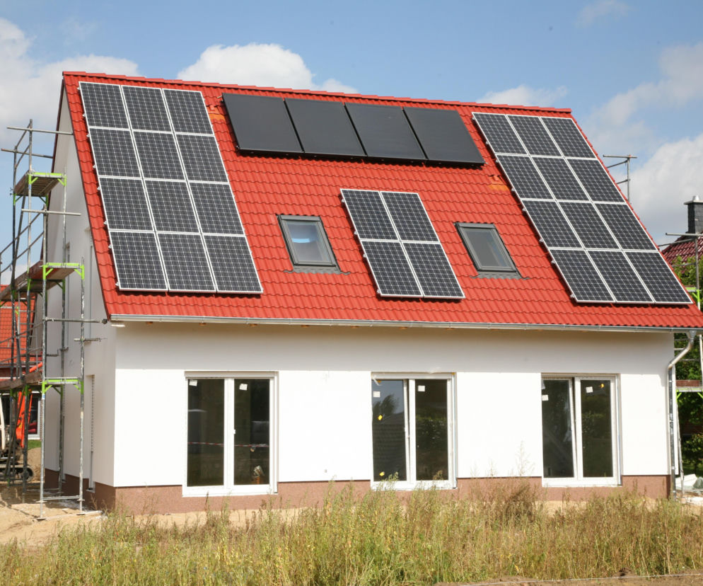 Dom jednorodzinny z czerwoną dachówką i panelami fotowoltaicznymi na dachu, symbolizujący program dopłat do energii odnawialnej. Szczegóły dotyczące dofinansowań na fotowoltaikę i magazyny energii znajdziesz na Super Biznes.