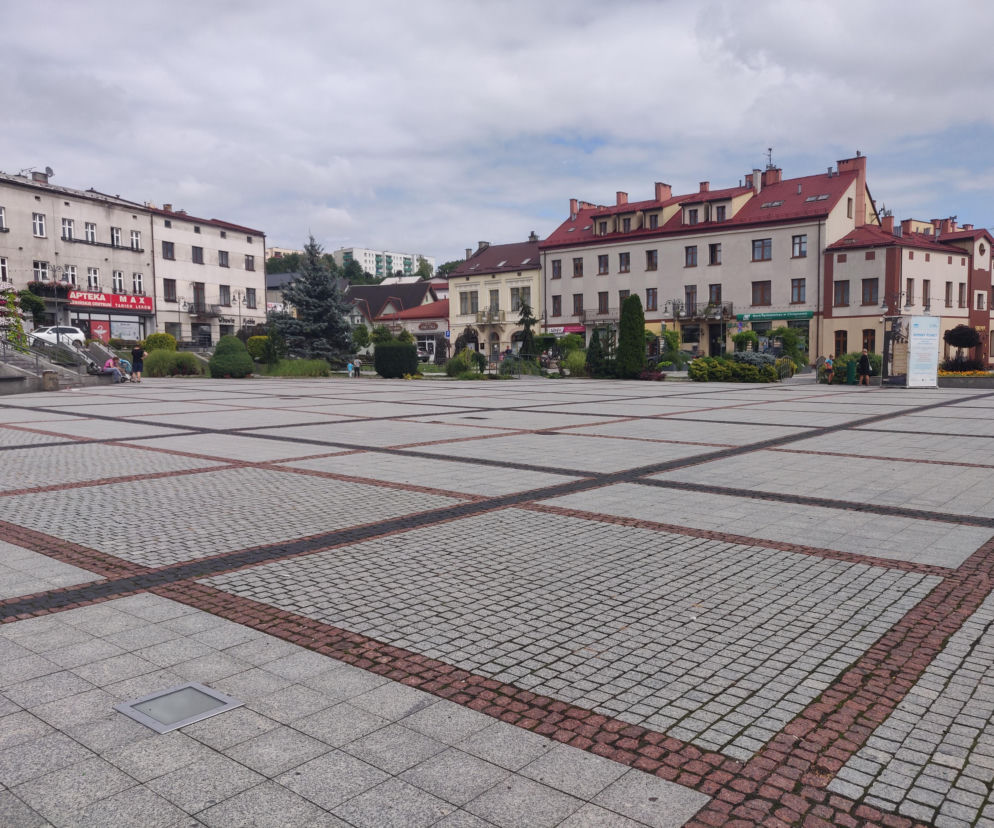 Rynek w Trzebini z zabytkowymi kamienicami o czerwonych dachach i brukowanym placem, otoczony zielenią i sklepami. Scena ilustruje piękno jednego z najstarszych miast Małopolski, o którym więcej przeczytasz na naszym portalu.